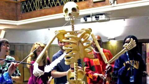 A group of five people animate a giant puppet depicting the skeleton of Thomas Paine