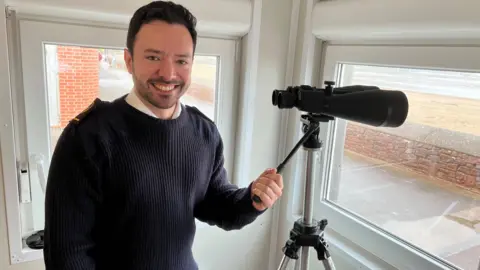 Josh is wearing a navy blue uniform and is smiling at the camera. He has short dark hair and is holding a pair of binoculars on a tripod.