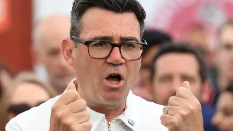 Andy Burnham is captured mid-gesture with both hands raised to chest level, his fingers curled into fists. He is wearing dark-framed glasses and a white zipped top. His mouth is open in mid speech.