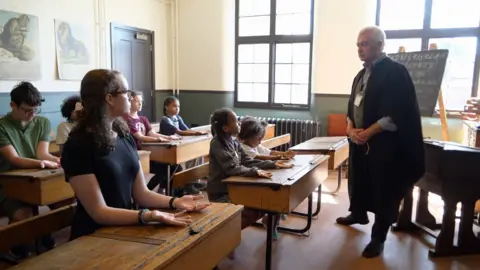 An old‑style classroom scene where students sit at wooden desks with their palms facing upward. The teacher stands at the front near a chalkboard filled with cursive alphabet practice.