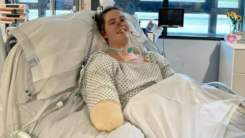 Lily McGarry Lily is lying in a hospital bed with medical tubes and a breathing support device, surrounded by monitors and equipment in a clinical room.