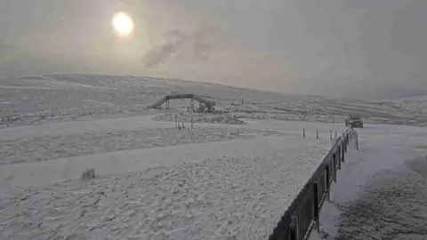 A webcam view of the road at the Bungalow looking towards Onchan. There is a metal footbridge in the distance and the ground is covered with snow.