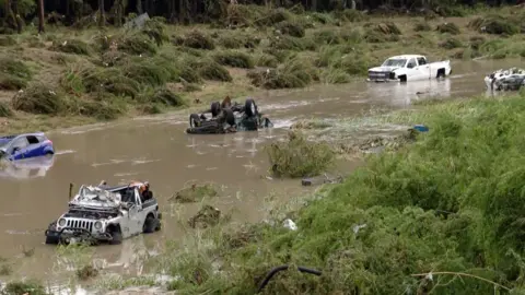Multiple damaged vehicles sit in a pool of water where flooding took over San Antonio. One is upside down and another is halfway submerged. Another appears to have flipped multiple times and its roof is damaged. 