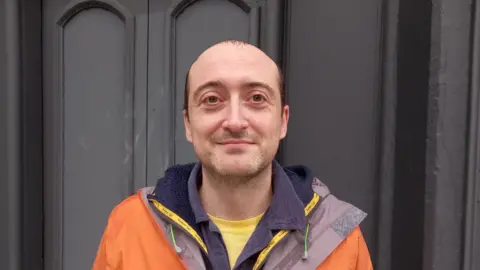 Darren John started the Save Station Street campaign after the Electric cinema shut. He has a mainly shaved head with some brown hair at his temples, stubble and wears an orange jacket with a grey lining over a blue top.