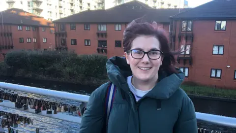 Sarah Hague seen with a canal behind her and housing in the background, is wearing glasses and an outdoor coat and carrying a bag. She is smiling and looks animated as she talks about energy prices.