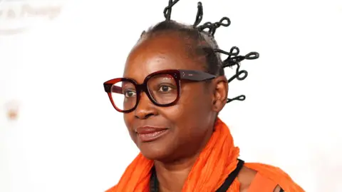 Koyo Kouoh wears thick rimmed glasses, an orange scarf and an orange top.