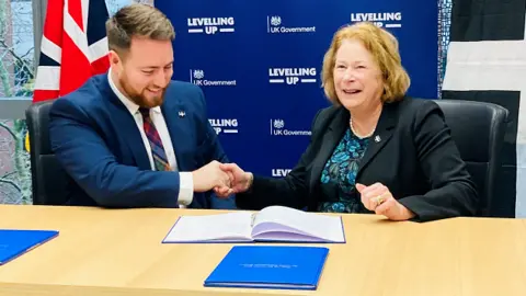 Cornwall Council A man and woman in suits shake hands, sat down at a table with an open treaty-style book in front of them.