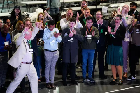 Jacob King/PA Wire A man in a cream suit with long white hair holds his arms aloft as a group of people wearing green rosettes cheer behind him 