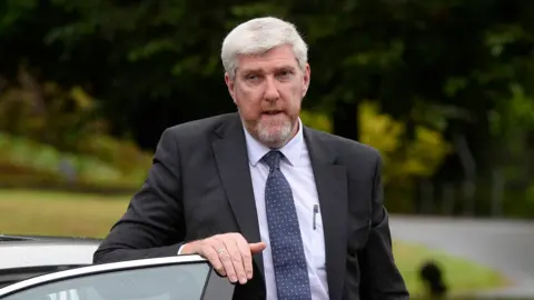 PA Media John O'Dowd, a man with grey hair and a ginger-grey beard and wearing a dark suit and blue tie