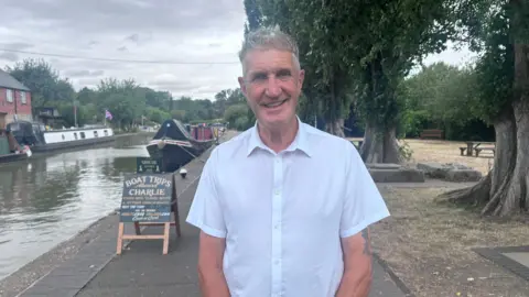 Kate Bradbrook/BBC A man with short grey hair and wearing a white short-sleeved collared shirt, is standing on a canal-side path next to an A-board sign advertising "BOAT TRIPS". There is a row of tall trees on the right of the path, with the water on the left. It has a number of barges moored on it. 