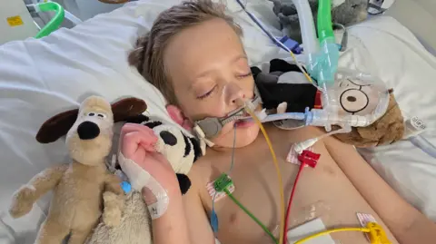A child lies in a hospital bed connected to multiple tubes and monitoring wires, including a breathing tube, with medical equipment surrounding the bed. Soft toys are tucked beside the child.