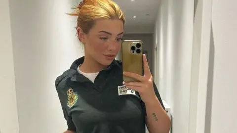 Lorna is using her mobile phone to take a selfie in a mirror here. She has blonde/ginger hair which is pinned up. She's wearing a green Welsh ambulance service polo shirt with a white shirt underneath. 