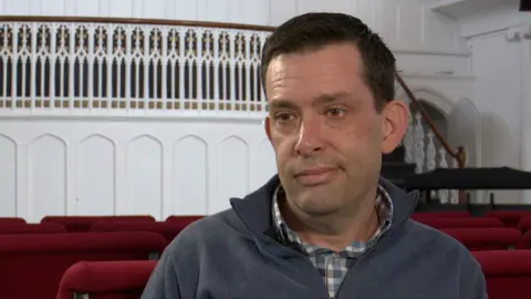 Pastor Drew Waller, mid 50's, in a room with rows of red chairs and an ornate white balcony behind them. They are wearing a collared shirt under a zip‑up sweater, positioned slightly off‑centre in the frame.