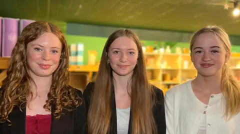 Darcy, Ailee and Hannah are standing side by side in a well-lit room with shelves filled with various items in the background. Darcy is on the left and has curly hair and is wearing a black blazer over a red top. Ailee is in the middle and has long straight hair and is wearing a black blazer over a light-colored top. Hannah is on the right and has blonde hair tied back and is wearing a white cardigan over a light-colored top