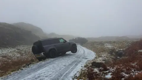 An icy single track road with two black cars run off it. The road is bordered by tall mountains.