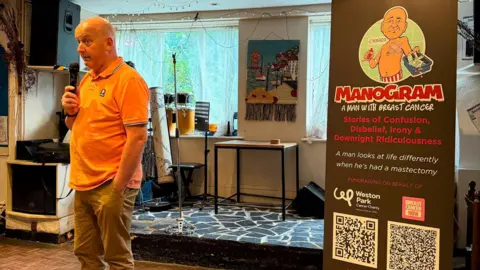 A bald man in an orange polo shirt talks into a hand held microphone