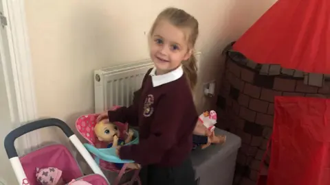 Lauren Marsh Bonnie Marsh is in the middle standing next to a toy highchair with a baby in it. She has long blonde hair in a ponytail and is standing next to a red indoor child's tent.