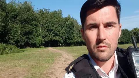 Handout photo issued by the Crown Prosecution Service of Ryan Connolly who is wearing a police uniform of white shirt and a black vest which has a communication device attached to it. He has straight dark hair, brushed into a side parting and has slight facial stubble. He looks directly into the camera, behind him a wide open space of grass is visible, with a path worn into it. Beyond that are trees in leaf and a bright blue sky