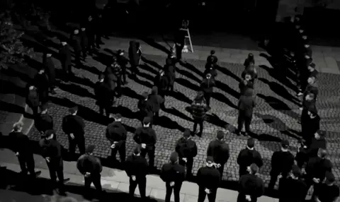 YouTube Men dressed in black, in military-style formation, stand in front of a picture of a teenager, Kriss Donald