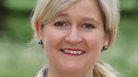 SNEE ICB Amanda Lyes looks at the camera and is wearing rounded metallic earrings and pink lipstick. She has blonde hair tiedback and the image shows her face close up