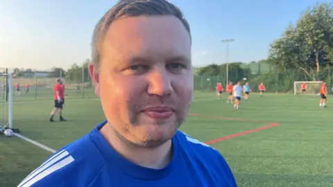A man in a blue sports shirt stands beside a football pitch, while players take part in a game in the background.
