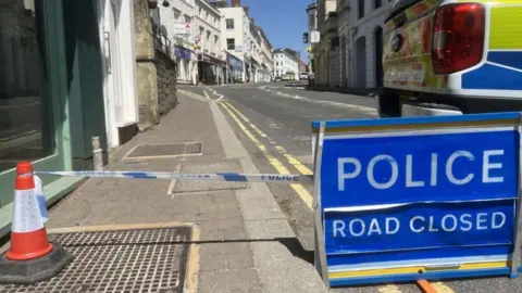 A blue sign which reads police road closed with blue and white tape tied to a traffic cone and an empty street behind