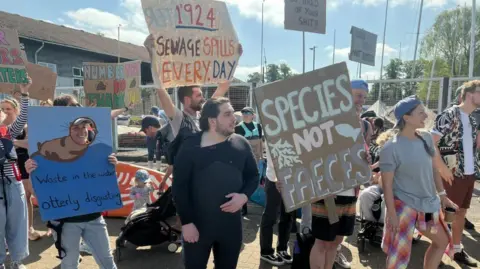 A group of people hold homemade signs. One reads "species not faeces", another says "waste in the water, otterly disgusting", while a third says "1'924 sewage spills every day". Other signs are visible but not legible. 