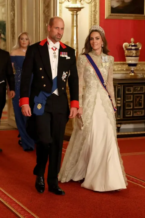 Fils Noble/Reuters Velsas princis atrodas tradicionālajā baltā kaklasaites apģērba kodā, un viņš ir dekorēts ar saviem karaliskajiem rīkojumiem un regālijām. Viņa jakai ir sarkanas atloka. Velsas princese nēsā britu dizainera Phillipa Lepley couture halātu ar pilna garuma, ar rokām izšautu zelta chantilly mežģīņu vakara mēteli virs zīda krepes halāta. Viņa valkā mīļāko mezglu Tiara.