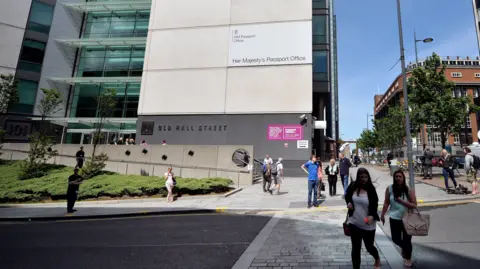 Stock image of people crossing the road in front of the Passport Office, which is a concrete and glass high-rise building