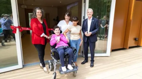Four people, including one wheelchair user, are at the entrance to a building, accessed through glass doors, with a wood panel floor. A red ribbon is tied across the front of the doors.