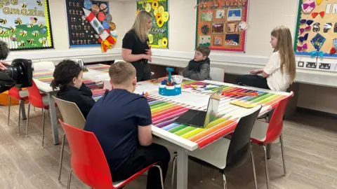 A woman with blonde hair stands beside a table with four children at it. The children are all looking at her and she is talking. There are brightly coloured sheets on the table. There are coloured paintings on the walls.