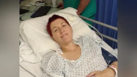 Cancer Research UK Ms Carey in a hospital bed wearing a clinical white-and-blue hospital gown. She has short red her and is smiling slightly at the camera. Her head is resting on a pillow and she has a paper wristband on her left arm which rests across her stomach. On her upper left arm is what appears to be a blue blood pressure machine. 
In the top right of the image, almost out of shot, is a medical professional at a computer wearing teal-coloured scrubs.