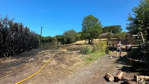 DSFRS A row of burnt out hedges and conifer trees can be seen to the left. A firefighter is walking away. Two people can be seen walking up a hill towards a nearby property on the right.