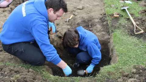 November Club  A man crouches down with a young boy who is digging with a small trowel in a small square area of earth 