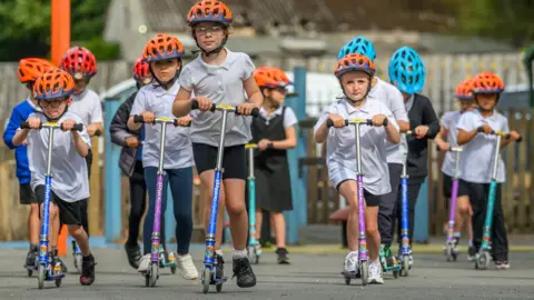 SBC A group of young school children on scooters with brightly coloured orange and blue helmets on