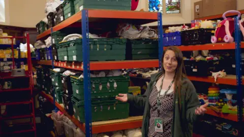 Tim John Becca Payne stands looking at the camera in front of shelves filled with crates of food
