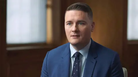 Wes Streeting, yang memiliki rambut cokelat dan mata biru, mengenakan setelan biru kekuatan udara dengan dasi berpola Angkatan Laut dan kemeja biru muda, duduk di kamar berpanel kayu gelap