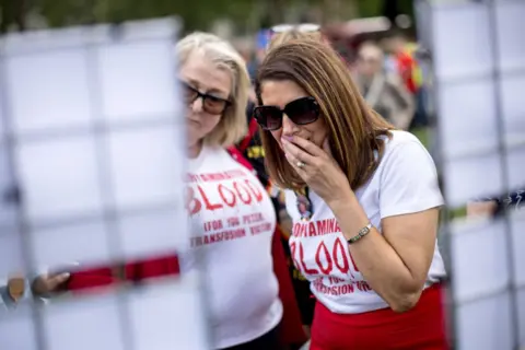 Getty Images Dua wanita berdiri di luar gedung di Westminster di mana laporan akhir tentang penyelidikan darah yang terinfeksi diterbitkan pada Mei 2020. Mereka berdua mengenakan T-shirt putih dengan slogan 'darah yang terkontaminasi" ditulis dengan tinta merah. Para wanita di sebelah kanan mengenakan kacamata hitam gelap dan memegang wajahnya ke mulutnya. 