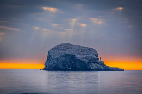 Asif Hanif The Bass Rock in the sea. The sky is dark with yellow light poking through the clouds. The horizon is a bright orange colour.