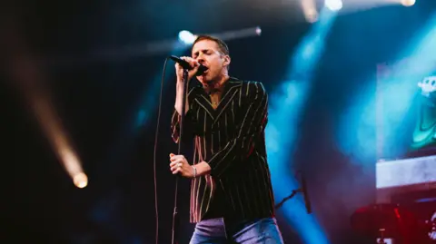 Nadine Ballantyne A male singer from the Kaiser Chiefs performing on stage. He is wearing blue jeans and a striped shirt. He has short dark hair and is standing in front of a microphone. His eyes are closed as he is singing.