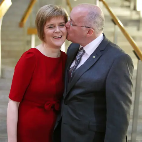 PA Media Nicola Sturgeon and Peter Murrell at the bottom of the staircase in the Scottish Parliament building. Sturgeon is wearing a red dress; Murrell is in a suit. He is kissing her on the cheek and she is smiling. 