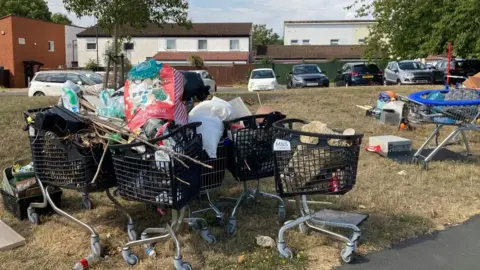 Several black shopping trolleys full waste sit on a patch of grass, there is housing and parked cars in the background. 
