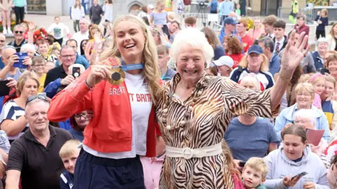 Pacemaker Hannah Scott wearing red Great Britain jacket is standing beside Lady Mary peters who is wearing a striped dress. Both are standing in front of a large crowd of people in Coleraine. 