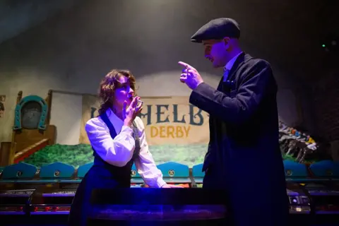 Joe Maher/Getty Images A petite woman with brown hair smokes a cigarette. The man opposite her is pointing at her as if he is speaking/gesturing. He is wearing a flat cap and a dark coat. 
