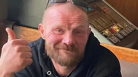 Greater Manchester Police Richard Crawt with short hair and light brown beard wearing a black hoodie and sunglasses on his head holding his thumb up and smiling inside a pub with a bandit machine behind him. 