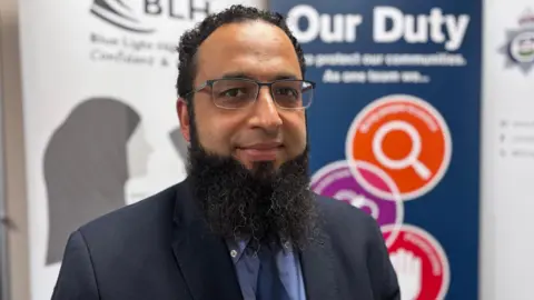 Det Sgt Yassin Desai, who has short dark hair, a beard and glasses, is wearing a dark suit with blue shirt and tie.