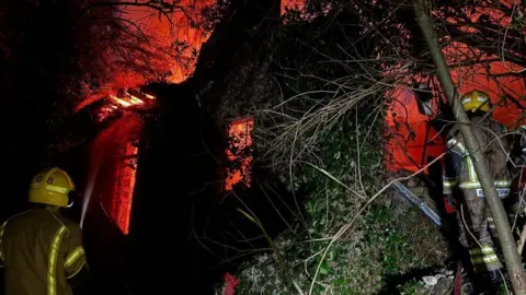 Backs of two firefighters with stone building in the background - it is engulfed by fire that is giving off a bright orange glow.