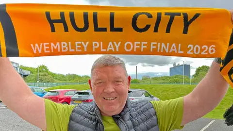 Linsey Smith / BBC Richard Heath holding up an amber and black Hull City Wembley play-off scarf.
He has short light hair and is wearing a lime green T-shirt and grey padded gilet 