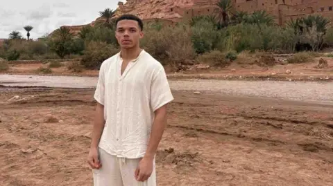Sam Rushton Young man wearing beige stands in the desert, posing for a photo.