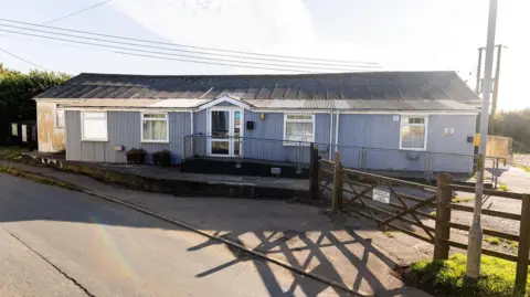 Cury Village Hall Committee Exterior of the single-storey property. The village hall has five windows and a glass entrance. A wooden gate leads into the property. It has an accessible slope  which leads to the entrance of the building.